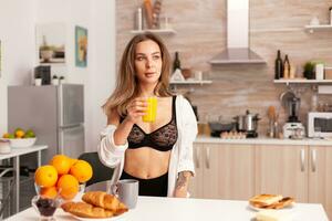 mujer participación vaso con Fresco naranja jugo durante desayuno vistiendo sexy negro lencería. joven sexy seductor sangre dama con tatuajes Bebiendo saludable, natural hecho en casa naranja jugo, refrescante domingo Mañana foto