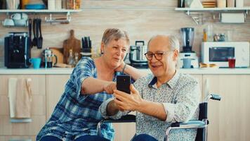 sonriente mayor mujer y su discapacitado marido en silla de ruedas utilizando teléfono inteligente en cocina. paralizado minusválido antiguo mayor hombre utilizando moderno comunicación tecnología. foto