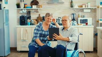 Senior couple laughing and waving during a video call with grandchildrens using tablet computer in kitchen. Paralysied handicapped old elderly man using modern communication techonolgy. photo