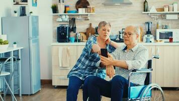 Senior woman and disabled husband in wheelchair surfing on internet using smartphone in kitchen. Paralysied handicapped old elderly man using modern communication techonolgy. photo