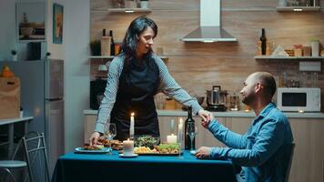 Encantado esposa servicio su marido. mujer preparando festivo cena con saludable alimento, Cocinando para su hombre un romántico cena, hablando, sentado a mesa en cocina, disfrutando el comida foto