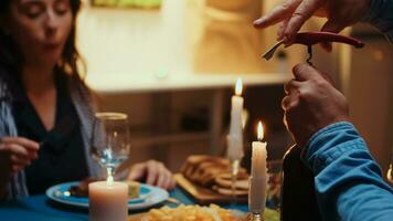 Close up of opening a bottle of wine during romantic dinner. Happy young couple sitting at the table in kitchen, talking, enjoying the meal, celebrating their anniversary in the dining room. photo