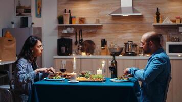 amantes comida juntos, comiendo y Bebiendo vino durante festivo cena en el cocina. contento Pareja hablando, sentado a mesa disfrutando comida a hogar teniendo romántico hora juntos sorpresa vela luces foto