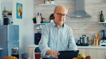 Elderly man surfing on internet using tablet computer during breakfast in kitchen. Elderly person with tablet portable pad PC in retirement age using mobile apps, modern internet online information technology with touchscreen photo