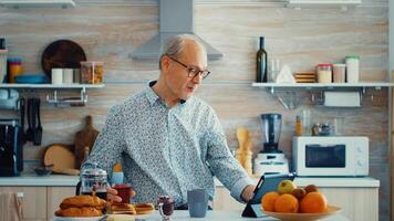 Senior man using tablet computer while drinking tea during breakfast in kitchen. Elderly person with tablet portable pad PC in retirement age using mobile apps, modern internet online information technology with touchscreen photo