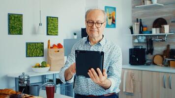 Happy older man in kitchen during breakfat using tablet pc. Elderly person with tablet portable pad PC in retirement age using mobile apps, modern internet online information technology with touchscreen photo