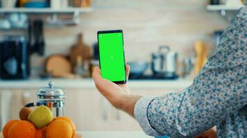 Mature man holding smartphone with green screen during breakfast in kitchen. Elderly person with chroma key isolated mockup mockup for easy replacement photo