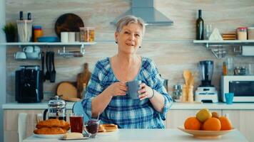 contento mayor mujer participación taza de café mientras mirando a cámara en ktichen durante desayuno. auténtico retrato de relajado mayor más viejo persona en el mañana, disfrutando Fresco calentar beber. sano sonriente adulto cara foto