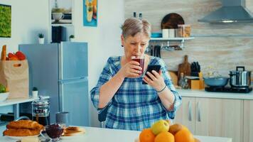 Senior woman using mobile gadget in the kitchen. Authentic elderly person using modern smartphone internet technology. Online communication connected to the world, senior leisure time with gadget at retirement age photo