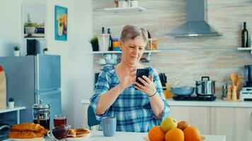 soltero mayor mujer surf en teléfono en cocina durante desayuno. auténtico mayor persona utilizando moderno teléfono inteligente Internet tecnología. en línea comunicación conectado a el mundo, mayor ocio hora con artilugio a Jubilación años foto