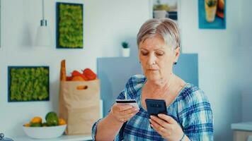 Senior woman paying online using phone app and holding credit card in kitchen during breakfast. Retired elderly person using internet payment home bank buying with modern technology photo