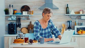 Senior woman searching online recipe using laptop in kitchen during breakfast. Elderly retired person working from home, telecommuting using remote internet job online communication on modern technology notebook photo