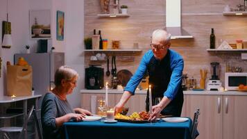 Jubilación maduro hombre surving cena a un comida habitación. mayor antiguo Pareja hablando, sentado a el mesa en cocina, disfrutando el comida, celebrando su aniversario con saludable alimento. foto