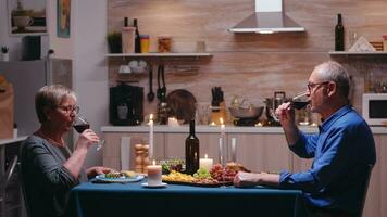 Elderly mature couple drinking a glass of wine during romantic dinner. Senior old people clinking, sitting at the table in kitchen, enjoying the meal, celebrating their anniversary in the dining room. photo