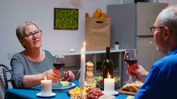 mayor relajado Pareja comiendo cena y Bebiendo lentes de rojo vino juntos en el cocina a hogar. anciano, retirado antiguo personas disfrutando el comida, celebrando su aniversario en el comida habitación. foto