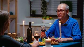 romántico mayor Pareja celebrando matrimonio a hogar, en el cocina sentado a el mesa, Bebiendo rojo vino y comiendo saludable alimento. hermosa mujer y hermoso hombre disfrutando gasto hora juntos. foto