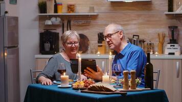 Envejecido, jubilado, Pareja teniendo un vídeo llamada en tableta durante romanítico cena. antiguo personas sentado a el mesa, hablando, utilizando el tecnología, Internet, celebrando su aniversario en el comida habitación. foto