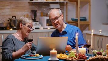 Envejecido, jubilado, Pareja compras en línea durante romanítico cena utilizando ordenador portátil. antiguo personas sentado a el mesa, hojeada, utilizando el tecnología, Internet, celebrando su aniversario en el comida habitación. foto