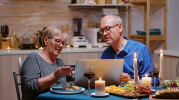 antiguo mujer pago en línea el fiesta utilizando ordenador portátil y tarjeta durante romántico cena. antiguo personas sentado a el mesa, hojeada, utilizando el tecnología, Internet, celebrando su aniversario. foto