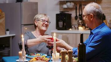 Mature man surprising woman with gift during fastive dinner sitting at the table in the kitchen. Happy cheerful elderly couple dining together at home, enjoying the meal, celebrating their anniversary photo