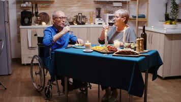 Paralyzed man toasting with wine with his wife. Senior old man in wheelchair dining with aged woman sitting in the kitchen. Imobilized paralyzed handicapped elderly husband having romantic dinner photo