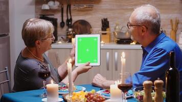 Old retired senior couple using green mockup screen digital isolated tablet computer. Aged people looking at green screen template chroma key display sitting at the table in kitchen during dinner. photo