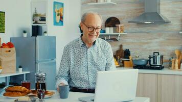 mayor hombre chateando y ondulación durante un vídeo llamada utilizando ordenador portátil en cocina participación un taza de café. mayor persona utilizando Internet en línea charla tecnología vídeo cámara web haciendo un vídeo llamada conexión cámara comunicación conferencia llamada foto