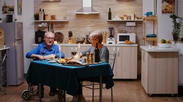 contento mayor mayor esposa comiendo romántico cena a hogar en el acogedor cocina con su discapacitado marido. inmovilizado paralizado minusválido mayor hombre teniendo romántico cena. foto