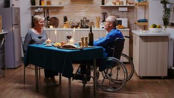 Senior couple coming home for romantic dinner. Old man in wheelchair dining with his cheerful wife sitting at the table in the kitchen. Imobilized paralyzed handicapped husband having romantic dinner photo