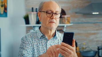 Old man surfing on social media using smartphone during breakfast in kitchen. Authentic portrait of retired senior enjoying modern internet online technology photo