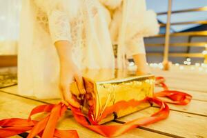 A woman in a white dress is holding a gold box with a red ribbon. She is wearing a crown on her head. The woman appears to be opening the gift box. photo