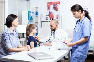 Pediatrician listening sick child heart using stethoscope in hospital office and nurse taking notes. Healthcare physician specialist in medicine providing health care services treatment examination. photo