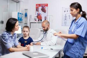 Doctor pediatry checking evolution of flu using stethoscope and nurse writing disease symptoms. Healthcare practitioner in medicine providing professional treatment services in hospital clinic. photo