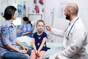 Checking body temperature of sick child in hospital room using digital thermometer. Healthcare physician specialist in medicine providing health care services treatment examination. photo