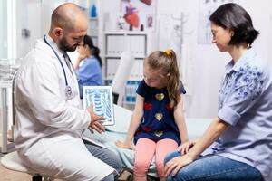 Specialist doctor holding tablet with radiography during medical examination of child in hospital office. Pediatrician explaining treatment illness providing healtcare services photo
