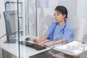 Focused nurse in blue coat checking appointments using computer for healthcare services treatment. Pediatrician assistant specialist in medicine providing professional healtcare services photo