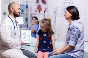 Pediatrician doctor explaining bones disease to mother using tablet with schelet image for sick child in hospital office. Physician specialist in pediatry medicine providing professional services photo