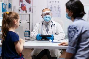 Doctor writing child diagnostic on tablet talking with mother for started treatment. Practitioner specialist in medicine providing health care services in new normal medical clinic photo