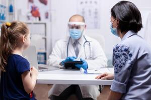 Pediatrician wearing face mask holding tablet pc while consulting child during clobal pandemic with coronavirus. Pediatrician specialist with protection mask providing health care service. photo