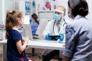 Pediatrician wearing face mask agasint coronavirus talking with child. Doctor specialist with protection mask providing health care service radiographic treatment examination. photo