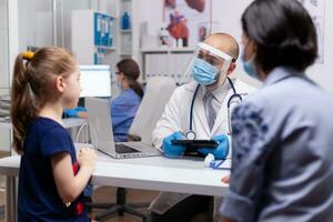 Pediatry specialist talking with little child about healtcare illness in medical clinic. Doctor in treatment disease wearing protective face mask against covid19 during global pandemic photo