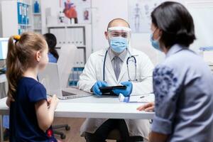 Doctor talking about child treatment with mother during coronavirus pandemic wearing face mask and holding tablet pc. Pediatrician specialist with protection mask providing health care. photo