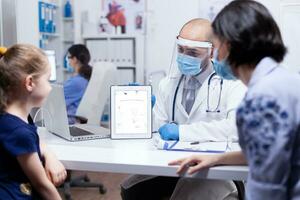 Pediatrician explaining skeleton to child and mother during consultation wearing fac mask against coronavirus. Pediatrician specialist providing health care service radiographic treatment examination photo