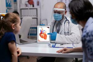 Pediatrician doctor holding digital tablet pc explaining heart condition in new normal medical clinic. Specialist physician talking about diagnostic resultes providing care services. photo