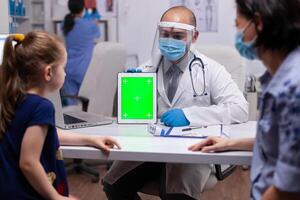 médico con cara máscara y visera utilizando verde pantalla tableta ordenador personal en examen oficina durante coronavirus pandemia. cuidado de la salud médico con croma llave cuaderno aislado Bosquejo pantalla foto