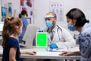 Therapeutist doctor using green screen tablet for examination in medical office. Pediatrician with protection gloves and mask against covid19 talking about treatment disease providing healtcare photo