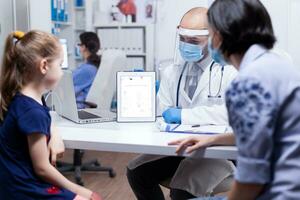 Pediatrician showing skeleton on tablet pc to mother and daugther wearing face mask agasint coronavirus during consultation. Doctor specialist providing health care service radiographic treatment examination photo