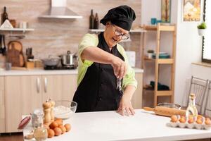 Cheerful senior lady making pizza in home kitchen using top flour for baking. Happy elderly chef with uniform sprinkling, sieving sifting raw ingredients by hand. photo