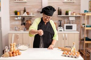 Senior lady chef smiling while preparing pizza sprinkling flour on kitchen table. Happy elderly chef with uniform sprinkling, sieving sifting raw ingredients by hand. photo