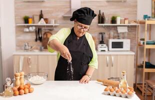 Preparation of delicious cookies in home kitchen by chef wearing apron. Happy elderly chef with uniform sprinkling, sieving sifting raw ingredients by hand. photo